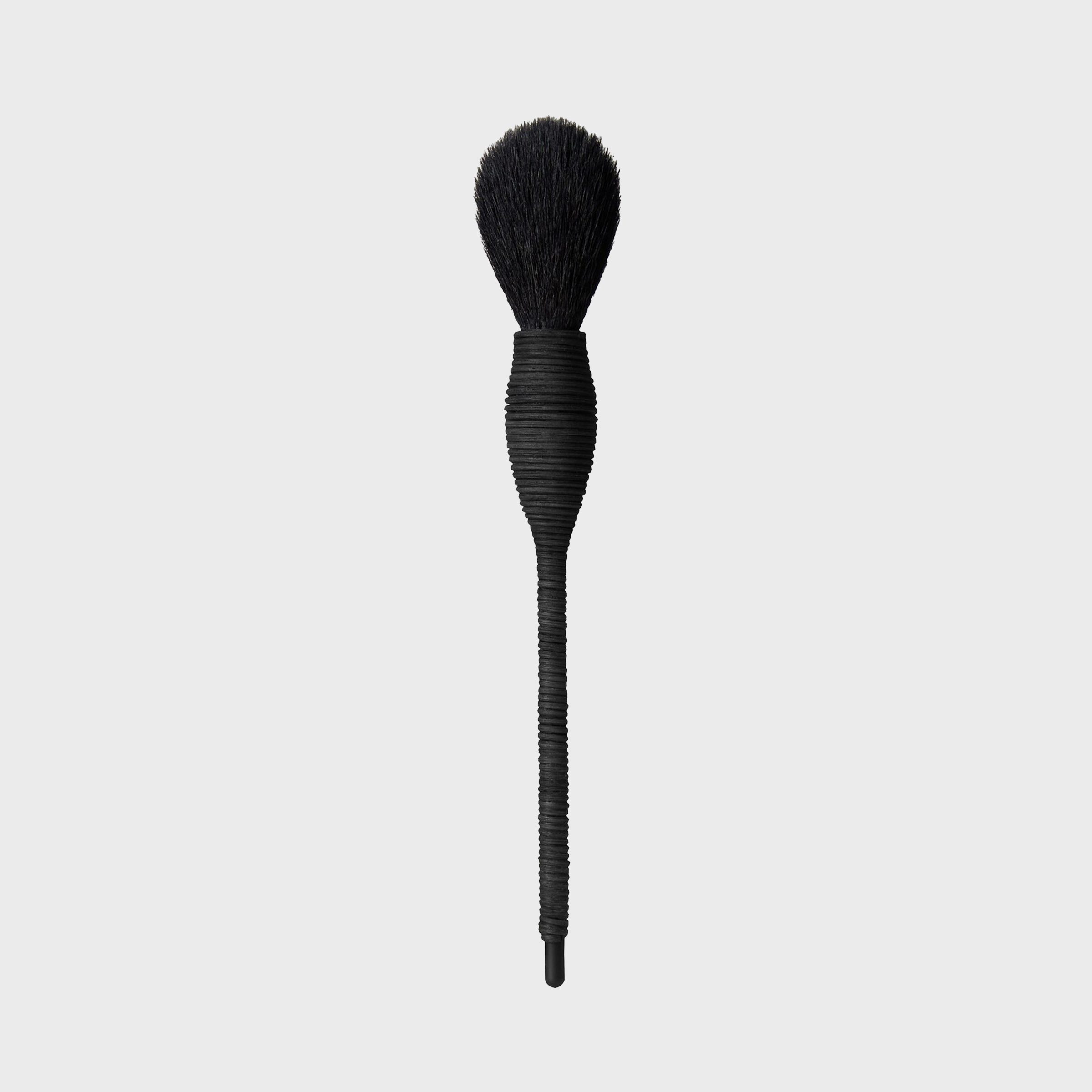 A black makeup brush with a textured handle stands upright against a plain gray background.