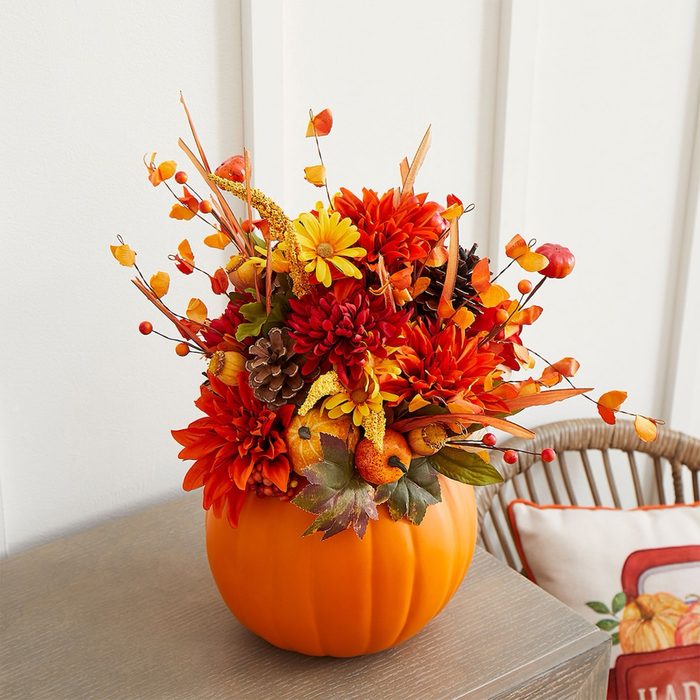 Pumpkin vase holds colorful autumn arrangement on wooden table. Rattan chair with festive cushion sits nearby against a light paneled wall.