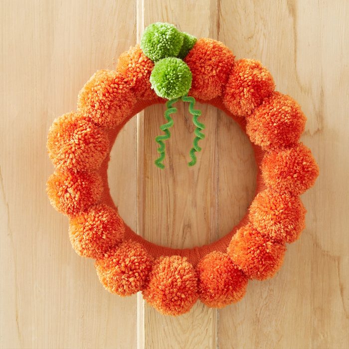 A pom-pom wreath hangs, displaying an orange circular design with green accents, against a wooden panel background.