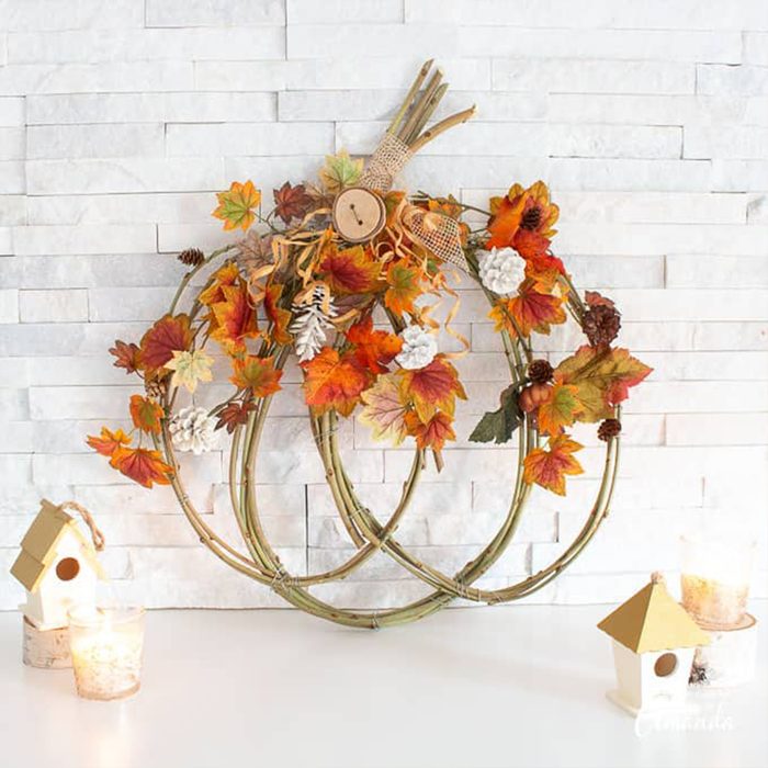 A wicker wreath decorated with autumn leaves sits on a white surface, flanked by candles and small birdhouses, against a brick background.