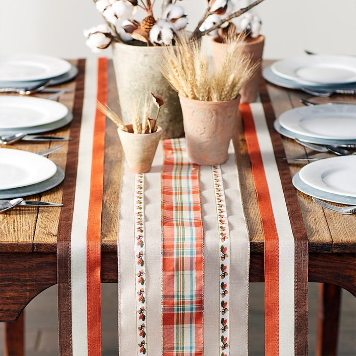 Table displays autumn-themed decorations with striped table runner; ceramic pots hold dried plants, and place settings include white plates and silverware.