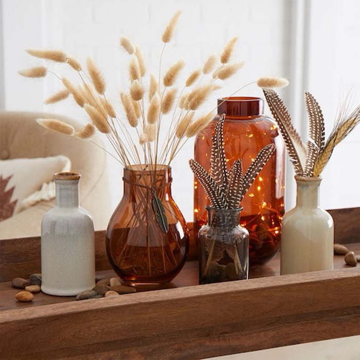 Vases display pampas grass and feathers, arranged on a wooden tray with scattered stones, in a bright, cozy room.
