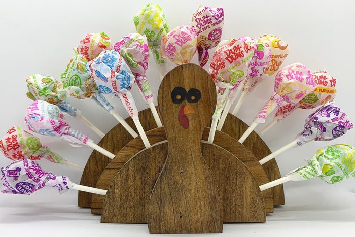 A wooden turkey holds colorful Dum Dums lollipops as feathers, arranged on a plain white background.