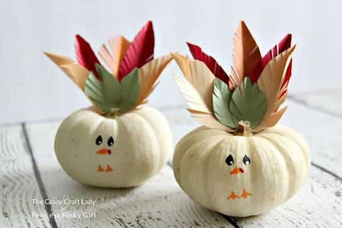 White pumpkins with drawn faces display paper feathers, resembling turkeys, on a wooden surface. 