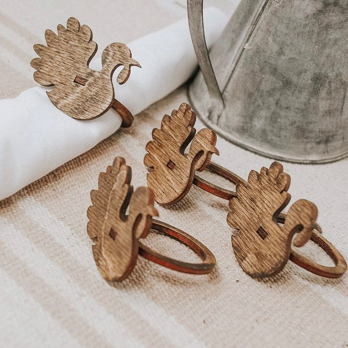 Wooden turkey-shaped napkin rings rest on a striped tablecloth, beside a metal pitcher, holding a white napkin in a rustic setting.