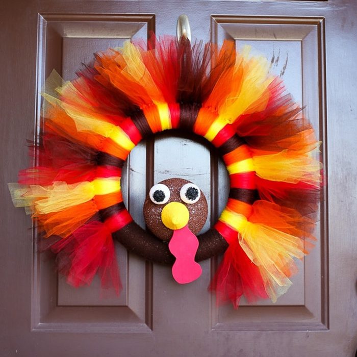 Wreath resembles a turkey with fabric feathers on a door, decorated in red, orange, and yellow hues, suggesting a festive, autumn theme.