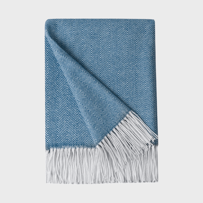 Blue herringbone-patterned blanket, partially folded, with white fringe, lying on a plain white background.