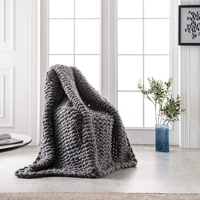 Chunky gray blanket draped on a chair in a bright, minimalist room with large windows, a small table, and a plant in a vase.