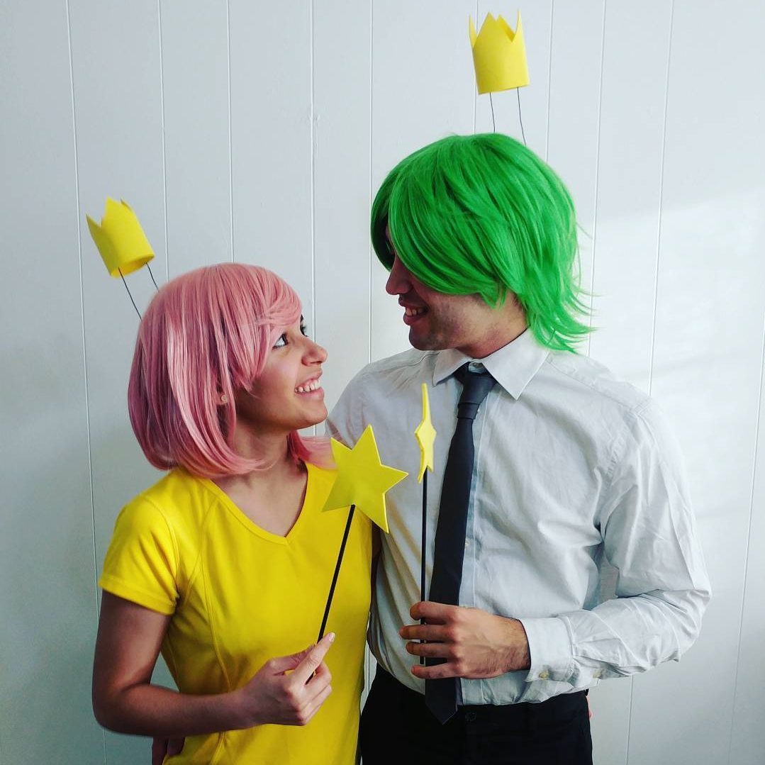 Two people wear colorful wigs and crowns, holding star wands, smiling at each other against a white wall background.