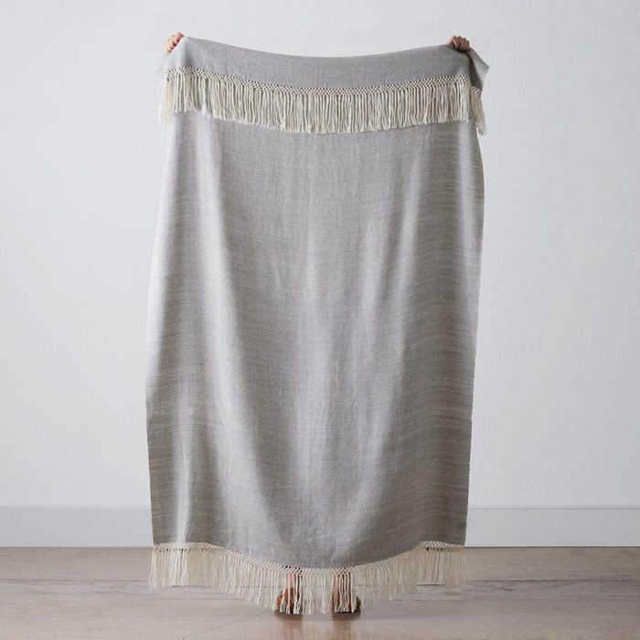 Blanket drapes vertically, held by hands, featuring fringe along the top and bottom, against a plain indoor background.