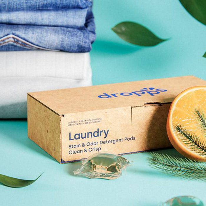 Box displays detergent pods beside stacked towels, sliced orange, and pine sprig on a light teal surface. Text: 