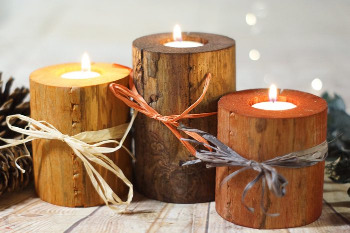 Three wooden candle holders with flames, tied with ribbons, sit on a wooden surface among pinecones and soft ambient lighting.
