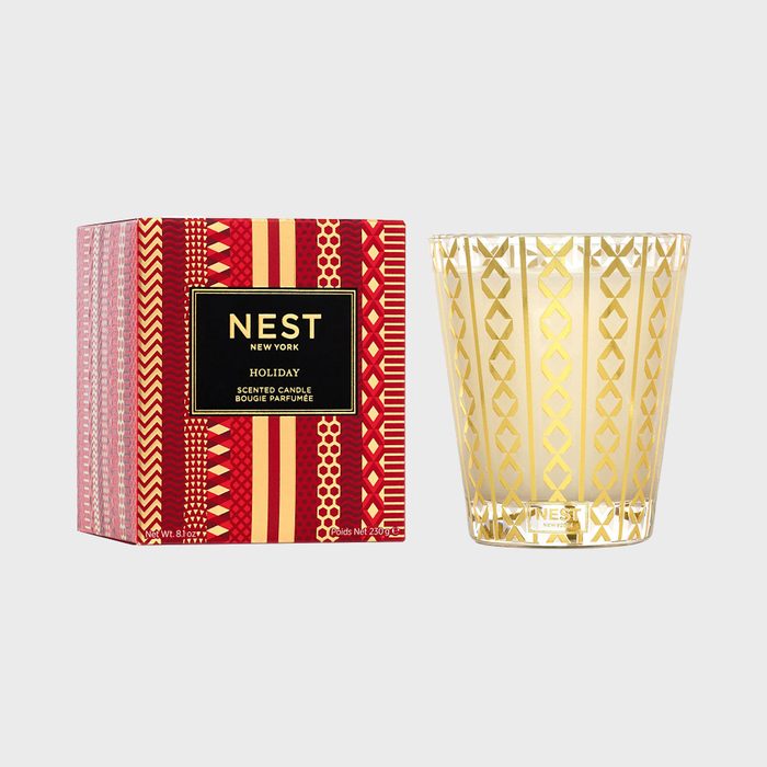 Candle sits beside patterned box, featuring NEST branding. Box displays festive red and gold designs, conveying a holiday theme.