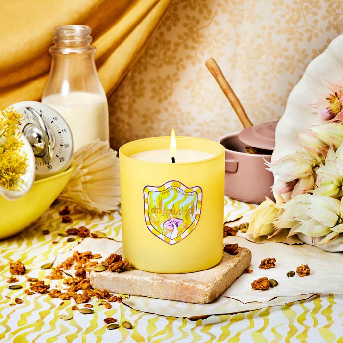 A yellow candle burns on a stone slab, surrounded by flowers and a milk jug, with a yellow patterned tablecloth underneath.