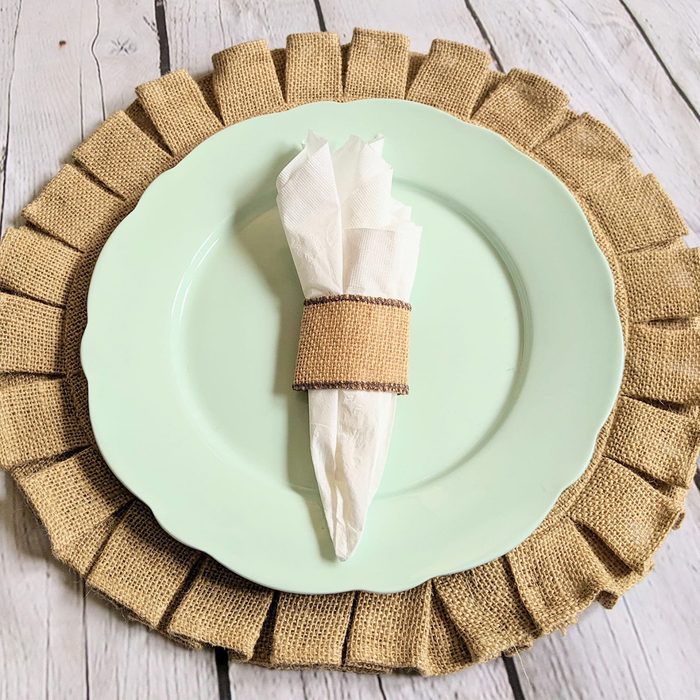 A green plate holds a napkin wrapped with burlap; placed on a wooden table with a burlap placemat.