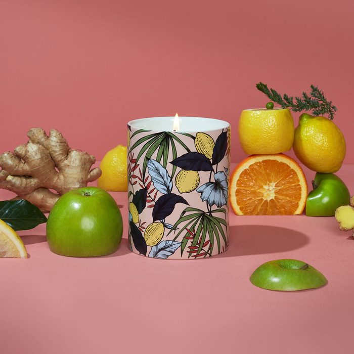 A floral-patterned candle burns surrounded by fruits and ginger on a pink surface.