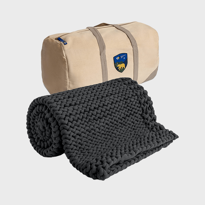 A rolled gray knitted blanket is next to a beige duffel bag with a colorful crest, set against a plain background.
