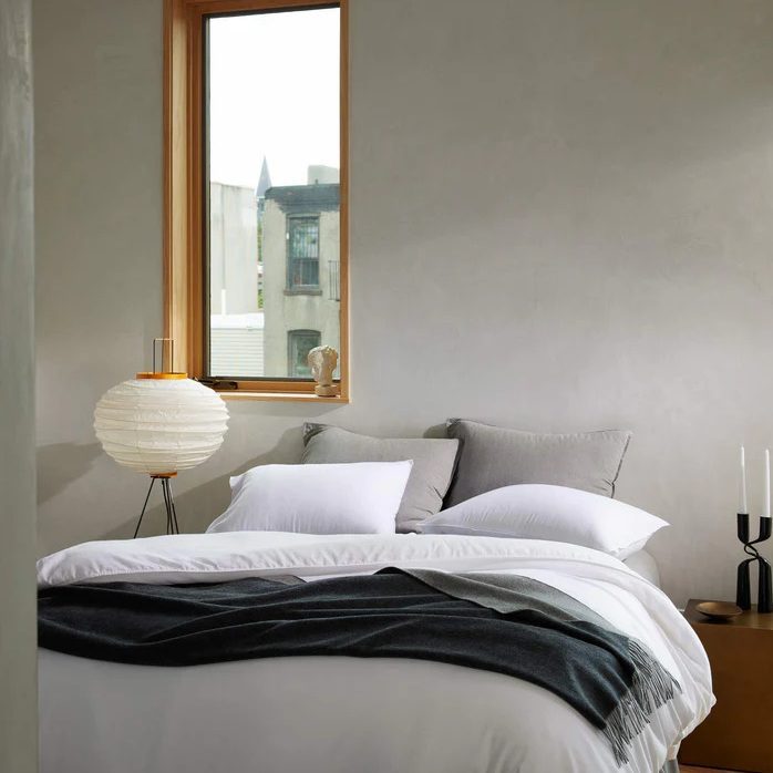 A neatly made bed rests beneath a window, flanked by a paper lantern and abstract candle holders, in a minimalist room.