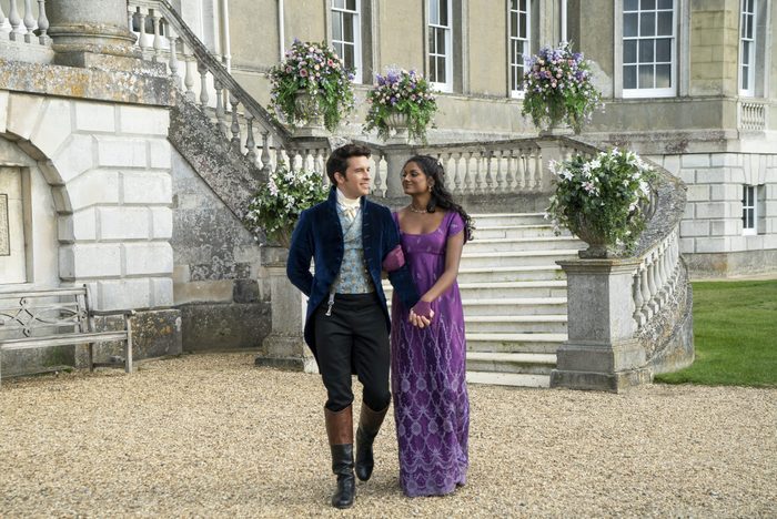 A couple walks arm-in-arm on gravel in front of a grand building, surrounded by stone steps and floral arrangements.