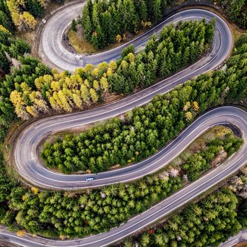 A winding road snakes through dense, colorful forest, viewed from above, with one car traveling along the curves.