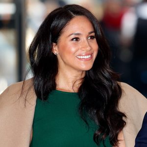 A person smiles, wearing a green top and tan coat, with long dark hair, indoors with blurred people in the background.