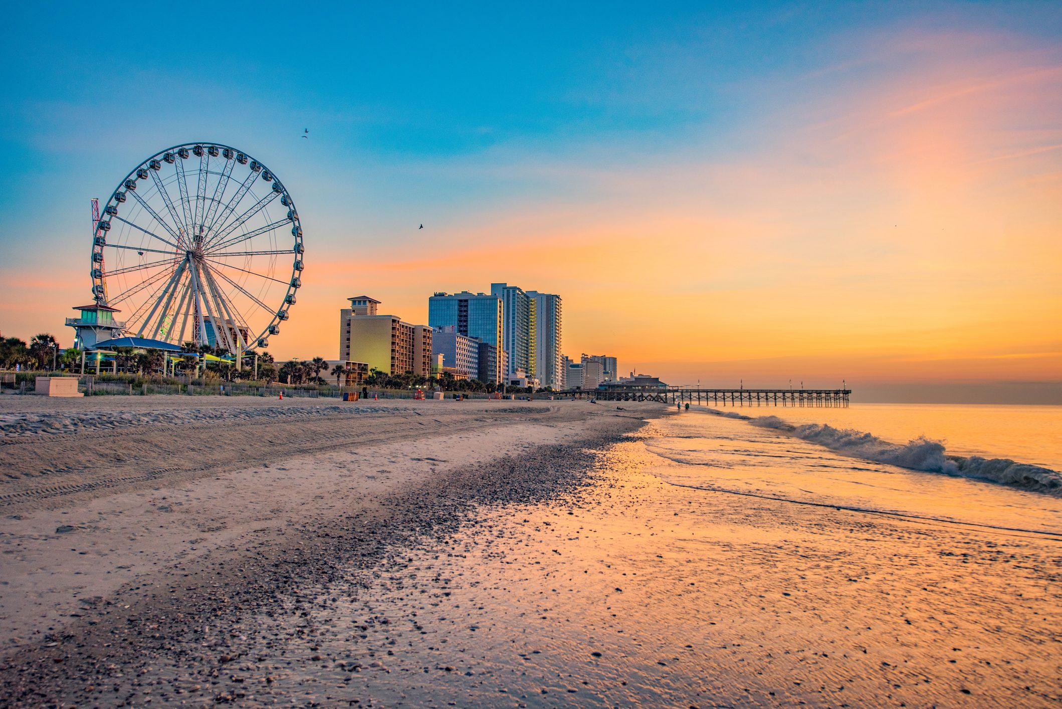 Ferris wheel stands tall beside high-rise buildings at a sandy beach during a vibrant sunset, with waves lapping the shore.