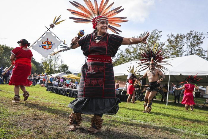 Dancer performs with feathered headdress, maraca in hand, during outdoor cultural festival with people and tents in background.