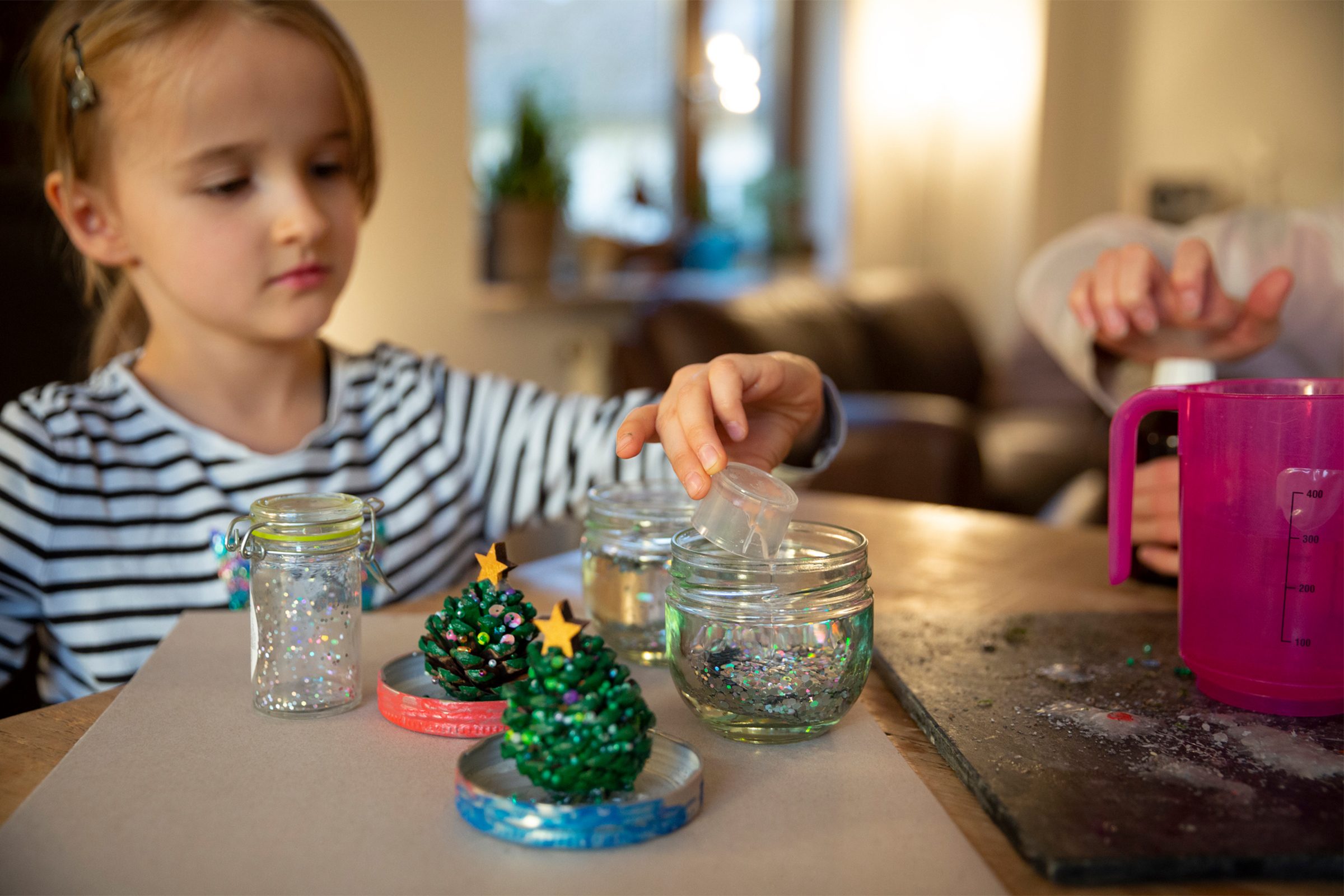 Children making Christmas snow globe decorations with glass jars, glitter and pine cones