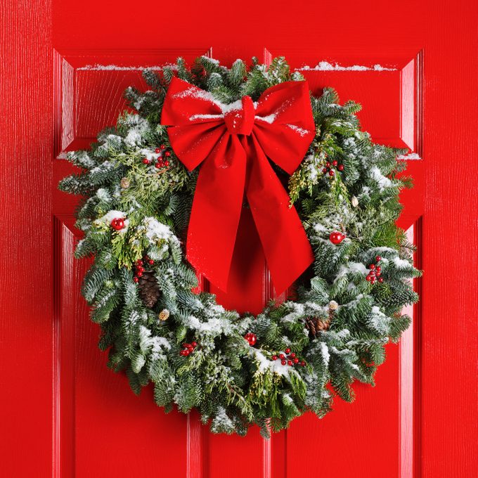 Wreath adorned with a large red bow hangs on a red door, dusted with snow, surrounded by green foliage and berries.