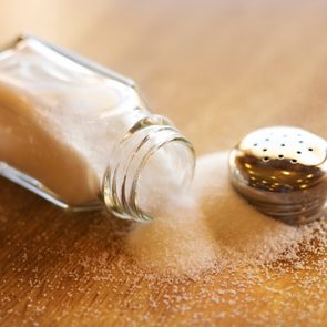 A tipped salt shaker spills granules onto a wooden table, scattering them across the surface.