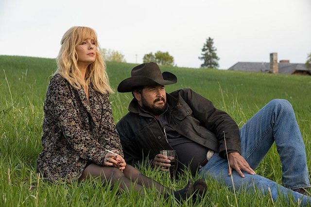 Two people sit on a grassy hill; one holds a drink. Rural setting with a distant building and trees under a clear sky.