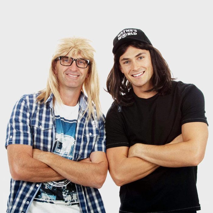 Two people wearing wigs and casual clothes stand smiling with arms crossed. White background with 