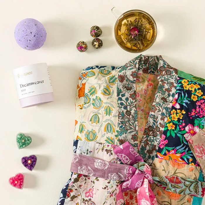 Colorful robe lies on a light surface, surrounded by a bath bomb, herbal tea, and floral chocolates, creating a spa-like atmosphere. Text: 