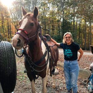 Horse stands with saddle, woman smiles beside it; sunlit forest backdrop.