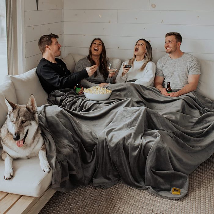 People sit under a grey blanket, laughing and drinking wine, on a white couch beside a large, relaxed dog, with popcorn in a bowl.