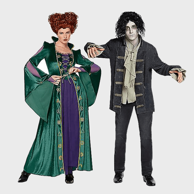 Two adults wear Halloween costumes; one as a witch in a green robe, the other as a zombie in tattered clothes. They're posing together.