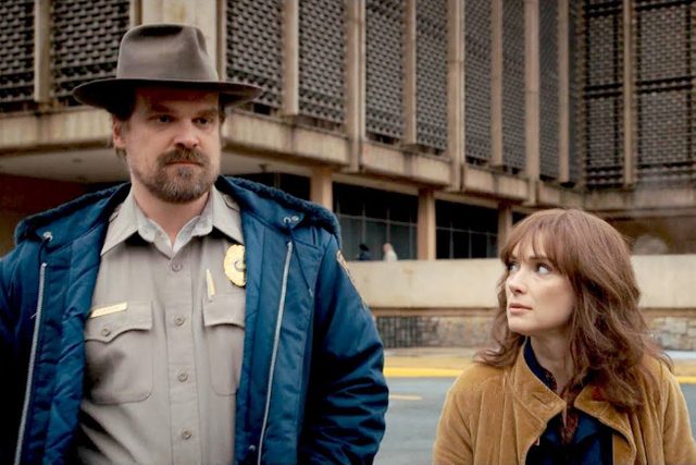 A man in a police uniform stands beside a woman, outside a large, concrete building.