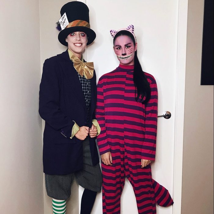 Two people in elaborate costumes stand in a room; one wears a top hat and bow tie, the other a striped cat outfit.