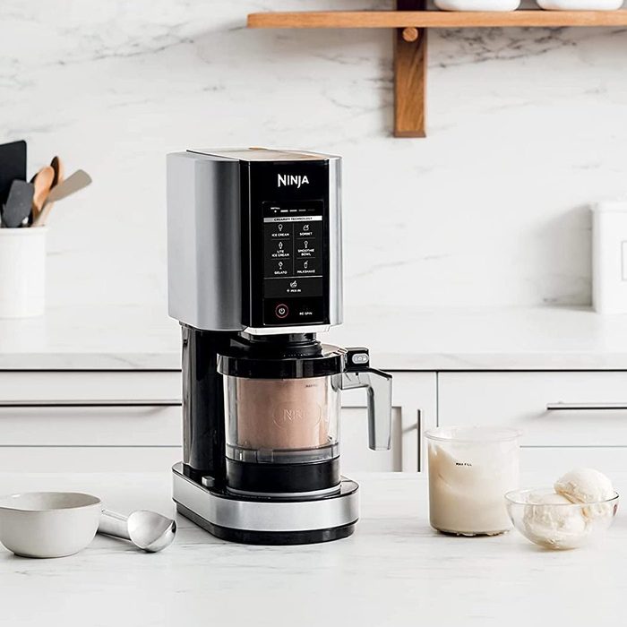 Ice cream maker sits idle on a white kitchen countertop, surrounded by a scoop, bowl, container of mixture, and a wall shelf above.