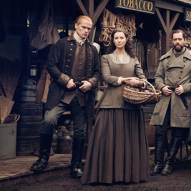 Three people stand in historical clothing; one holds a basket. They stand in front of a shop with a 
