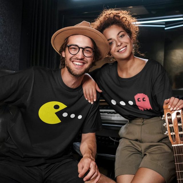 Two people smiling in a music studio, wearing Pac-Man themed shirts, one with a yellow Pac-Man and another with a pink ghost.