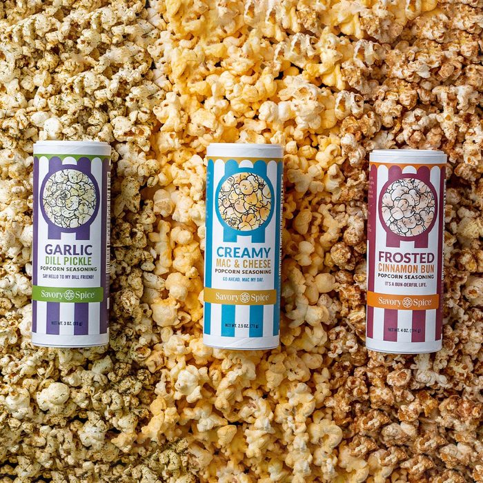 Seasoning containers stand upright, surrounded by corresponding flavored popcorn varieties. Labels read: 