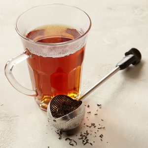 Glass mug holds steaming tea; metal infuser with loose tea leaves rests nearby on a light surface.