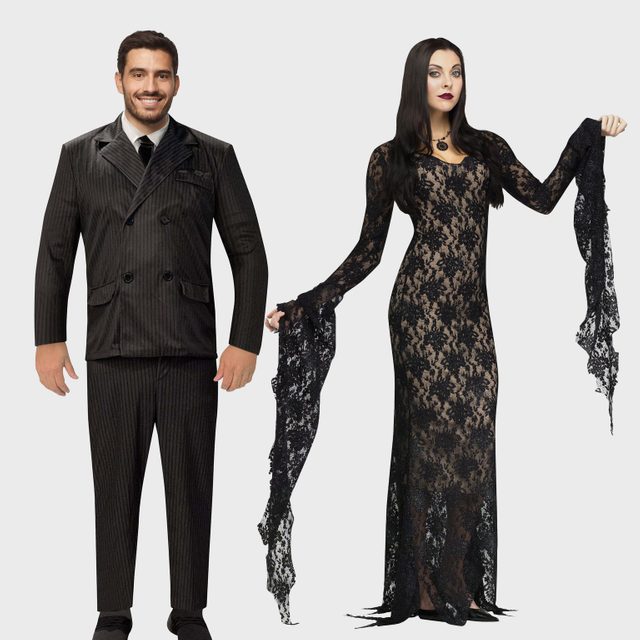 Two people wearing gothic costumes pose together, one in a pinstriped suit, the other in a black lace dress against a plain background.
