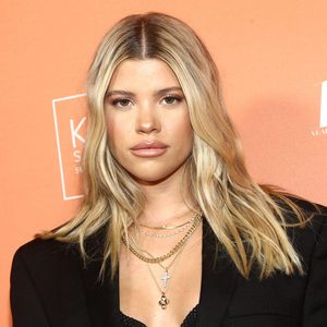 Person posing against orange backdrop, wearing layered gold necklaces and a black top; background features indistinct white text and logos.