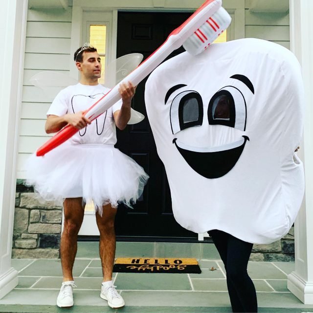 Fairy in tutu wields giant toothbrush near smiling tooth costume; standing on porch with welcome mat reading 