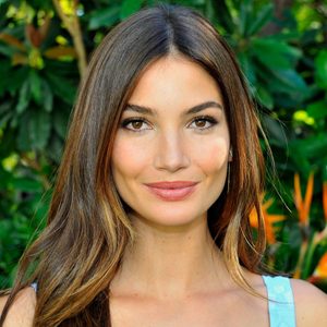 A woman smiles subtly, standing against a backdrop of lush green foliage.
