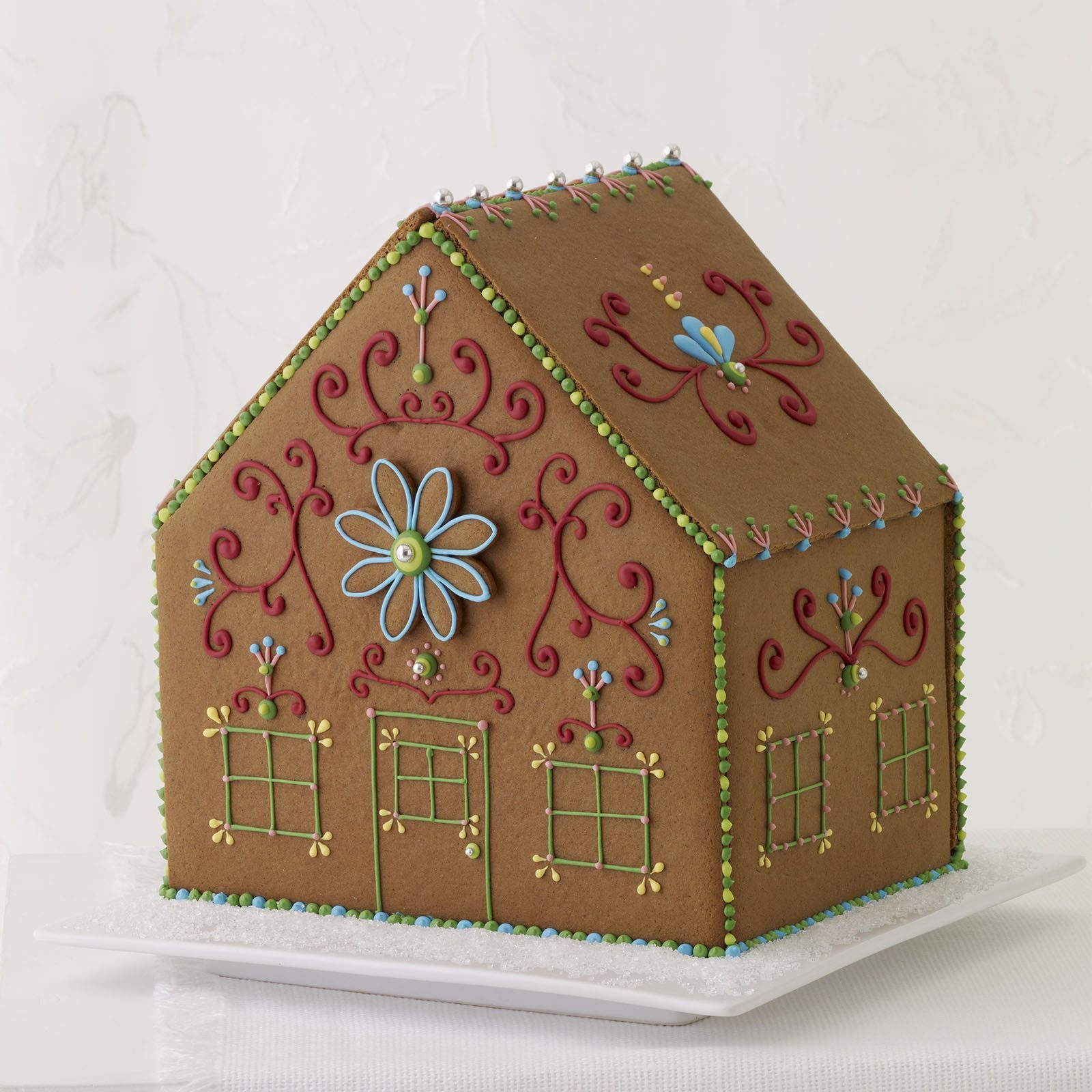 Gingerbread house stands decorated with colorful icing patterns, resting on a white plate amidst a simple, light-toned background.