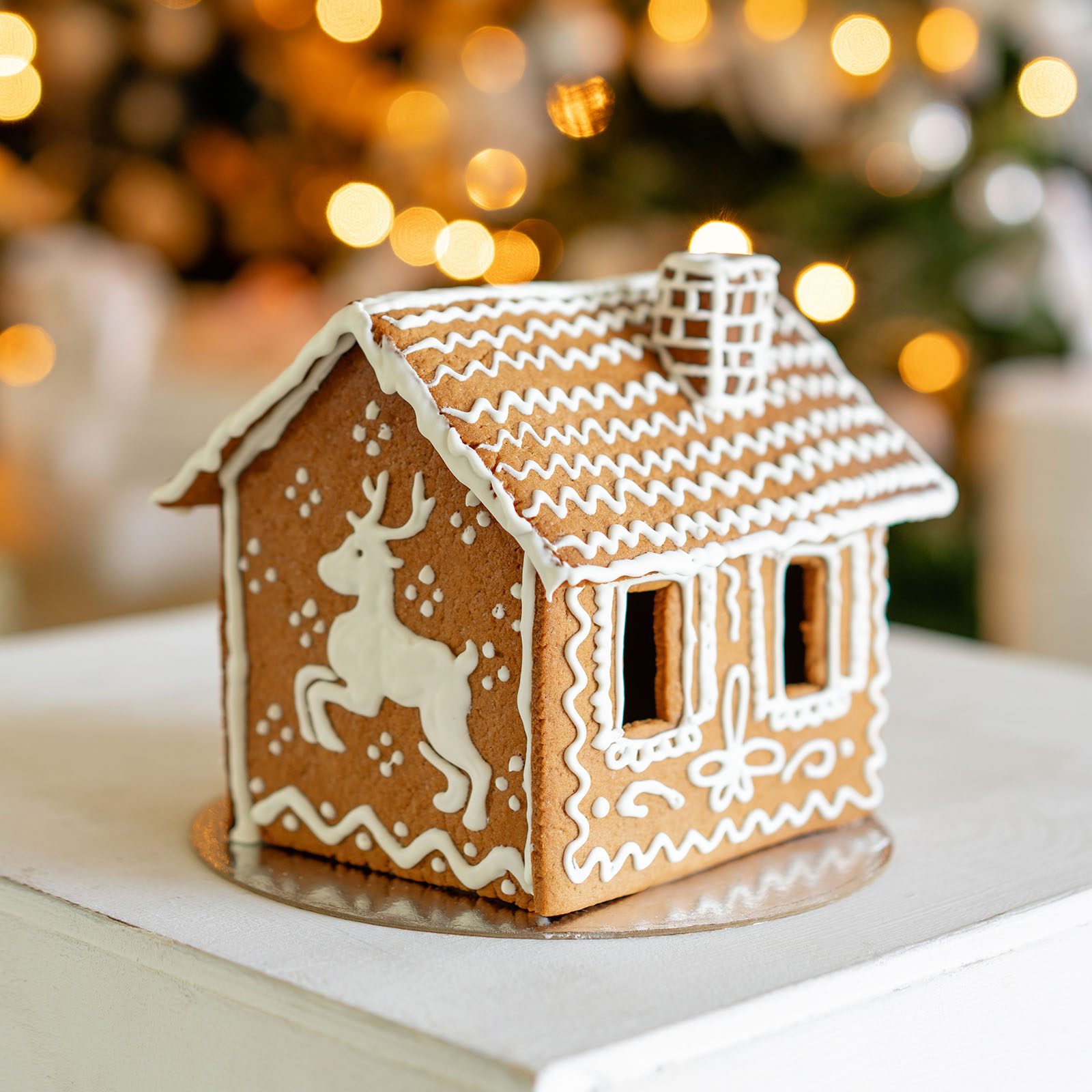 Gingerbread house sits adorned with white icing, featuring reindeer design, set on a table against a blurred background of warm holiday lights.
