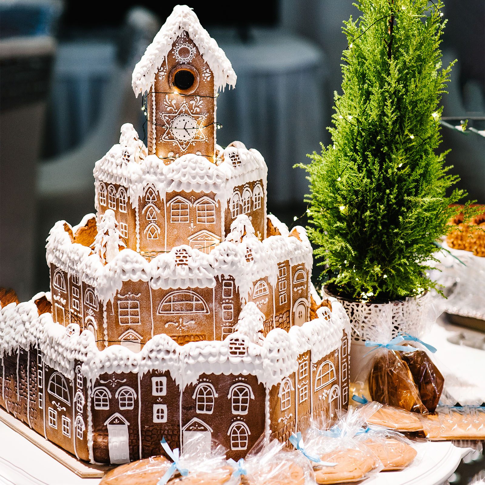 Gingerbread house towers intricately decorated with white icing, standing on a table next to a potted evergreen tree and wrapped cookies.